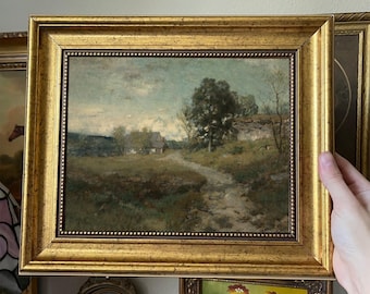 May include: A framed landscape painting with a gold ornate frame. The artwork depicts a dirt path leading to a small house, with trees and a cloudy sky in the background. The painting has a vintage aesthetic.