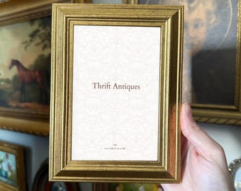 May include: A rectangular picture frame with a metallic gold finish and a stepped, beveled edge design. The center insert features a white background with a faint damask pattern, the text Thrift Antiques, and the dimensions 10.2 cm x 15.2 cm.