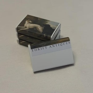 May include: A stack of matchboxes with a vintage painting of a white dog on the top. The matchbox in the foreground has the text "THRIFT ANTIQUES" printed on it.