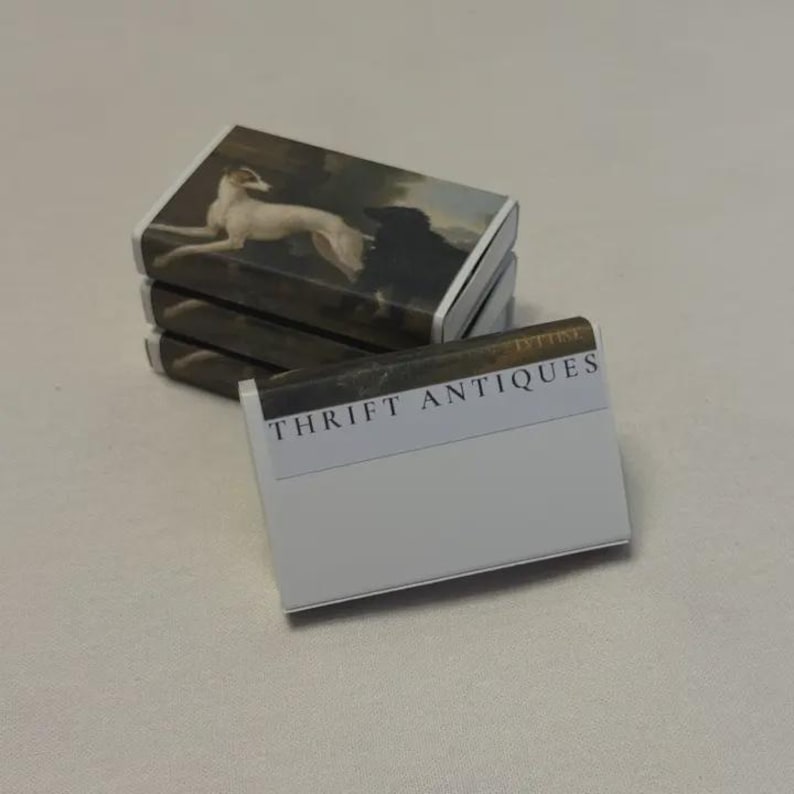 May include: A stack of matchboxes with a vintage painting of a white dog on the top. The matchbox in the foreground has the text "THRIFT ANTIQUES" printed on it.