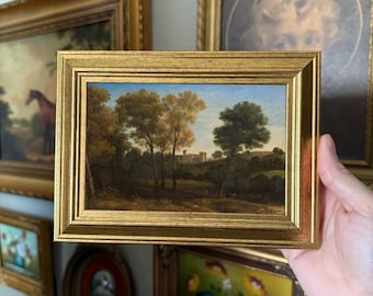 May include: A framed oil painting depicting a landscape scene with a castle in the distance. The painting features a lush green forest with trees and a blue sky with white clouds. The frame is gold and ornate.