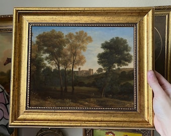May include: A framed landscape painting featuring a castle in the distance, trees, and a field. The painting is in a gold ornate frame. The artwork has a blue sky and a variety of green and brown trees.