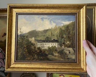 May include: A framed oil painting depicting a landscape scene with a white building in the distance, surrounded by trees and a cloudy sky.