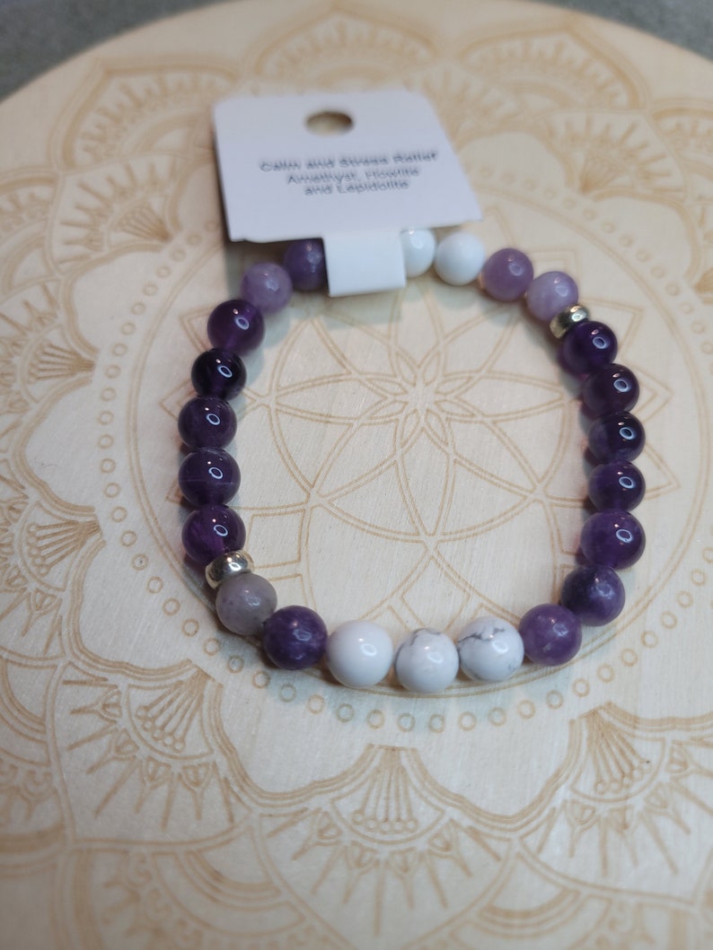 May include: A purple, white, and gray gemstone bracelet with a tag that reads "Calm and Stress Buster Amethyst, Howlite and Lepidolite".