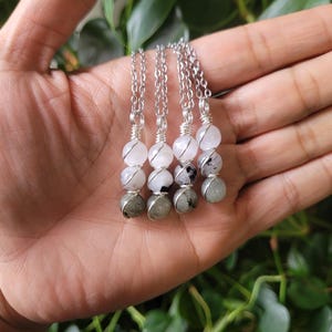 May include: Four silver chain necklaces, each featuring a combination of white and gray gemstone beads. The necklaces are held in a hand, with a backdrop of green foliage. The gemstones are wire-wrapped and strung together.