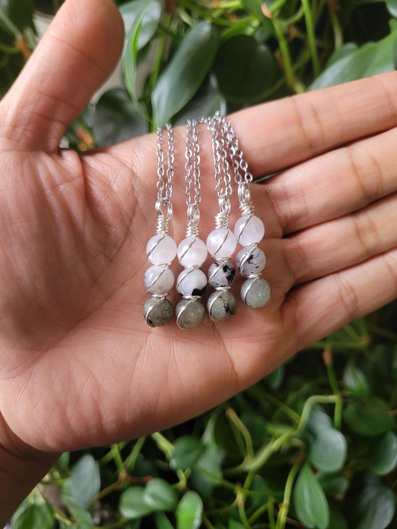 May include: Four silver chain necklaces, each featuring a combination of white and gray gemstone beads. The necklaces are held in a hand, with a backdrop of green foliage. The gemstones are wire-wrapped and strung together.