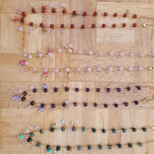 May include: Four beaded bracelets in shades of red, pink, purple, and green, each with a gold chain and clasp. The bracelets are adorned with various charms and crystal accents, displayed on a wooden surface.