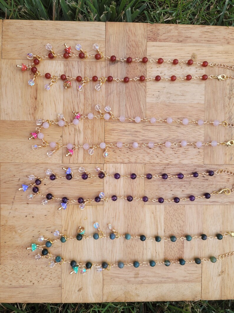 May include: Four beaded bracelets in shades of red, pink, purple, and green, each with a gold chain and clasp. The bracelets are adorned with various charms and crystal accents, displayed on a wooden surface.