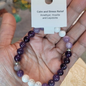 May include: A purple, white, and gray beaded bracelet with silver accents. The bracelet is labeled "Calm and Stress Relief Amethyst, Howlite and Lepidolite".