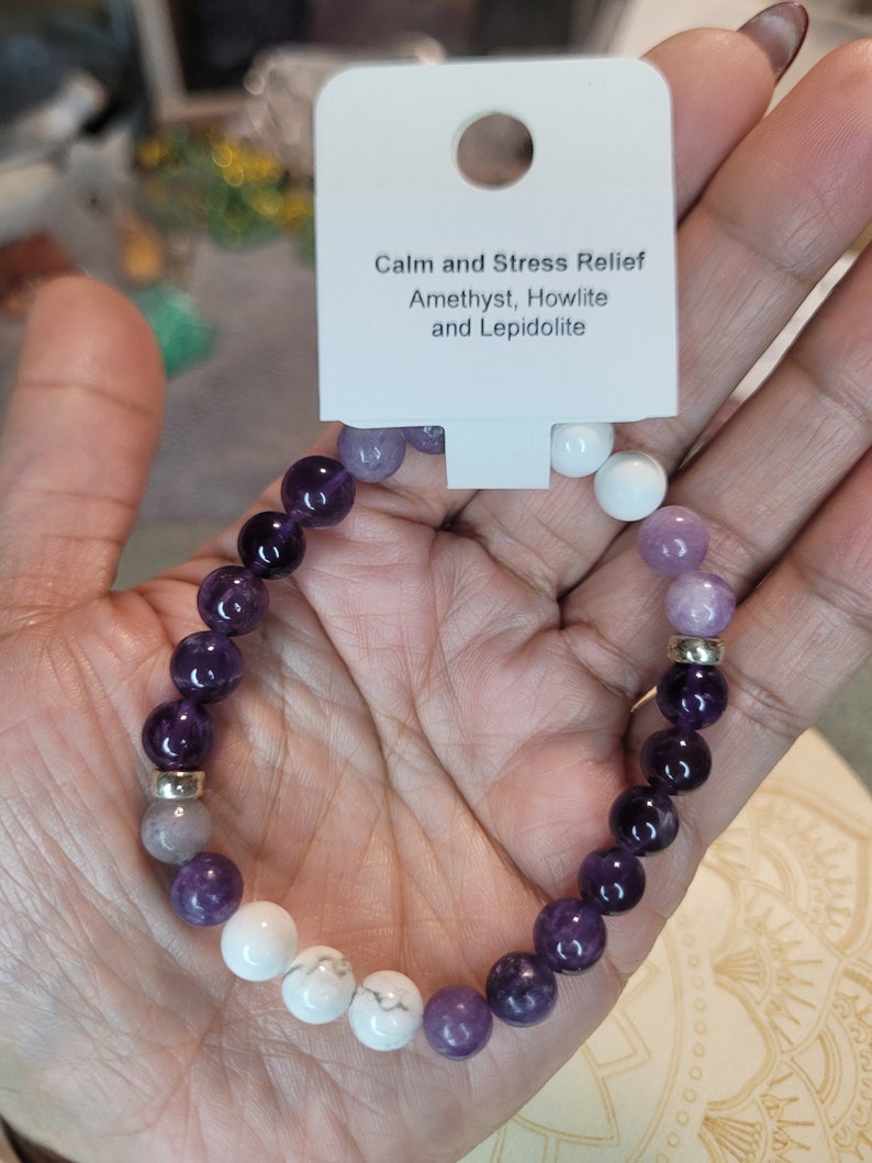 May include: A purple, white, and gray beaded bracelet with silver accents. The bracelet is labeled "Calm and Stress Relief Amethyst, Howlite and Lepidolite".