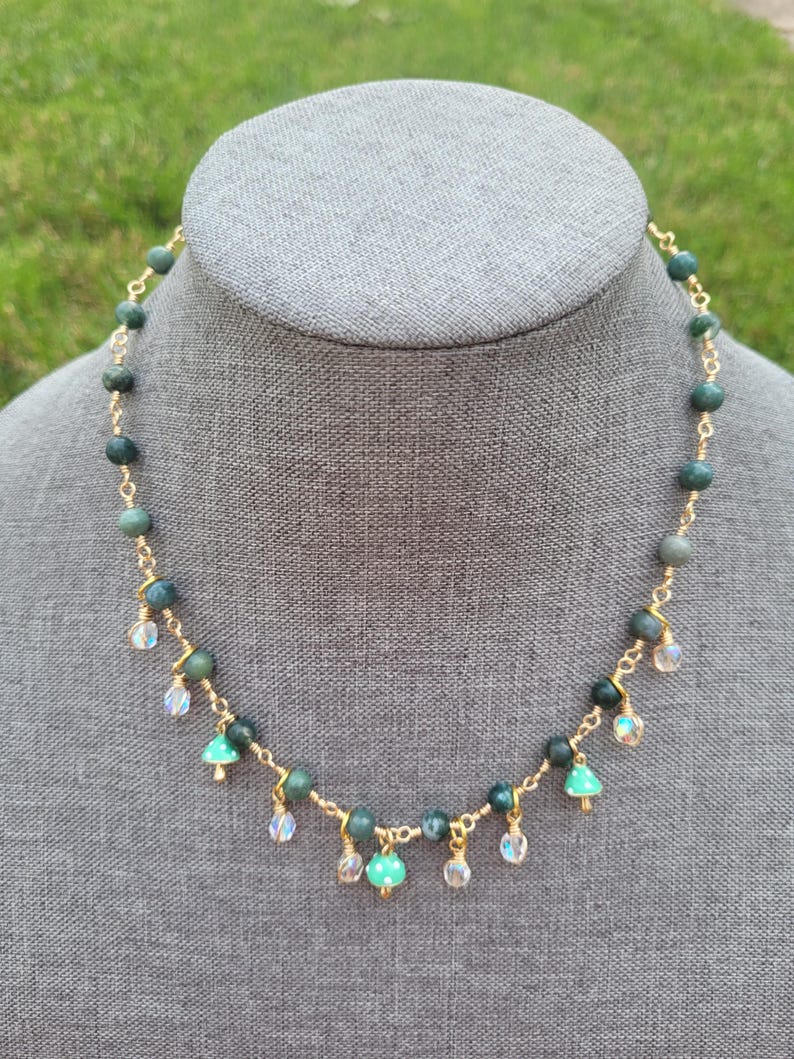 May include: A gold-toned necklace with green and clear beads. The necklace features a series of round, dark green beads connected by gold wire. Small, clear, and green beads dangle from the necklace, creating a delicate, eye-catching design.