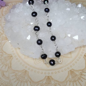 May include: A delicate silver chain bracelet with black pearl beads. The bracelet is displayed on a white crystal surface.