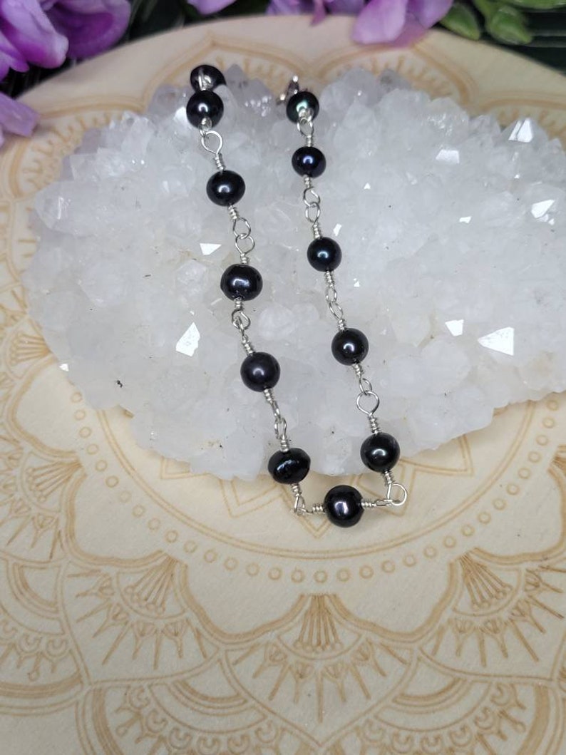 May include: A delicate silver chain bracelet with black pearl beads. The bracelet is displayed on a white crystal surface.