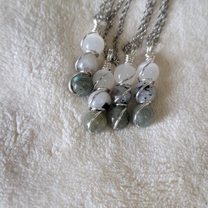 May include: Four silver chain necklaces, each featuring a series of three gemstone beads. The beads are white, gray, and iridescent green, wrapped in silver wire. The necklaces are displayed on a white, textured surface.