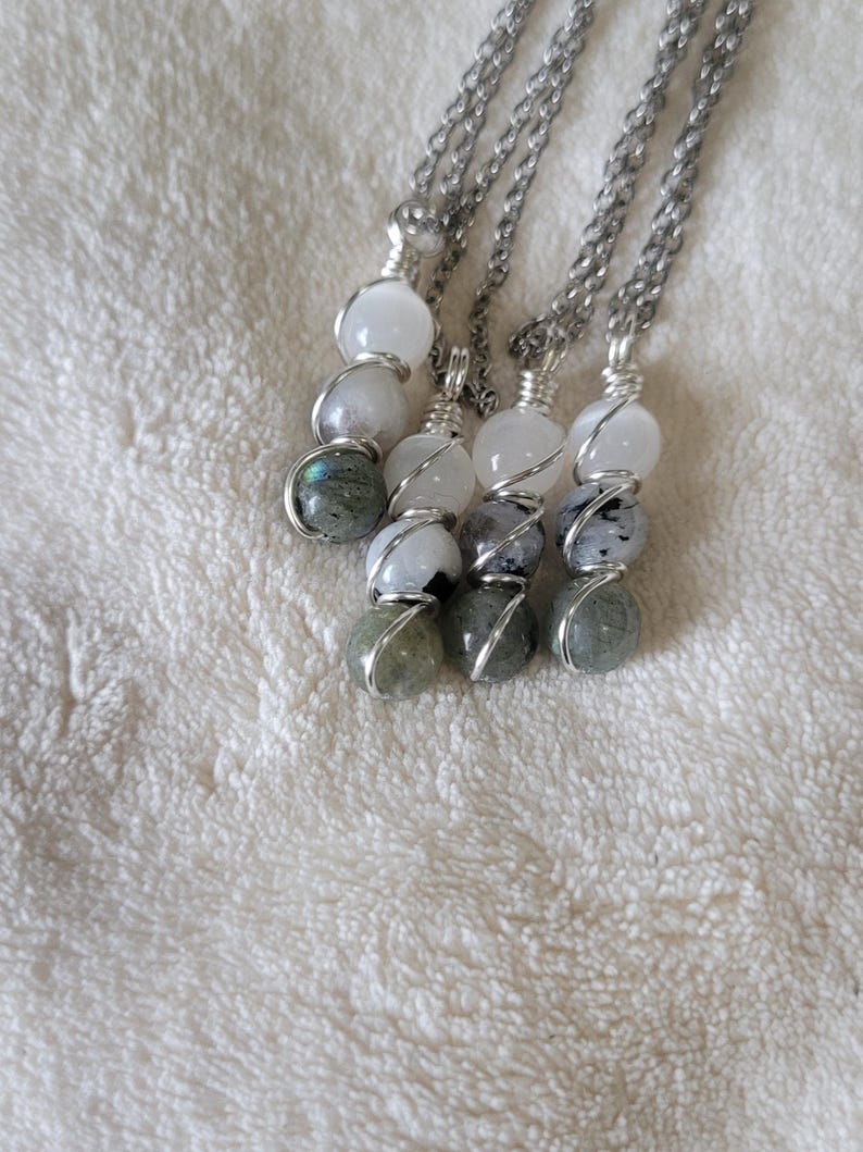 May include: Four silver chain necklaces, each featuring a series of three gemstone beads. The beads are white, gray, and iridescent green, wrapped in silver wire. The necklaces are displayed on a white, textured surface.