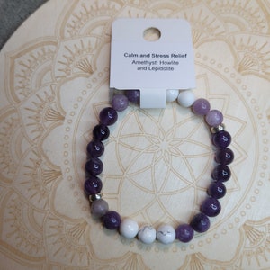 May include: A purple, white, and gray beaded bracelet with a silver charm. The bracelet is on a wooden surface with a mandala design. The tag attached to the bracelet reads "Calm and Stress Relief Amethyst, Howlite and Lepidolite."