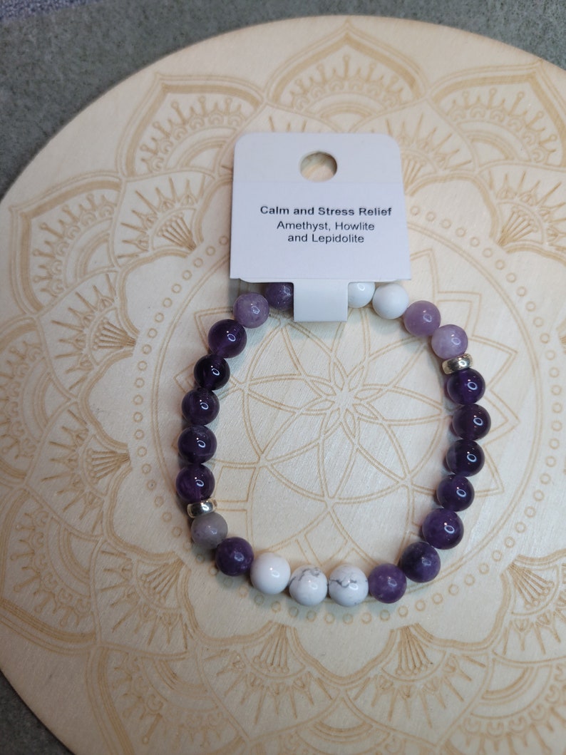 May include: A purple, white, and gray beaded bracelet with a silver charm. The bracelet is on a wooden surface with a mandala design. The tag attached to the bracelet reads "Calm and Stress Relief Amethyst, Howlite and Lepidolite."