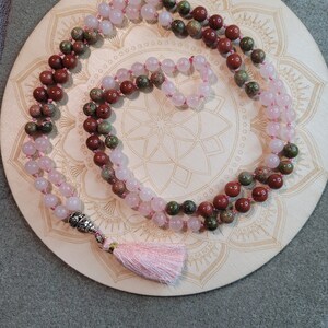 May include: A pink, green, and red gemstone mala with a silver tassel. The mala is on a wooden mandala.