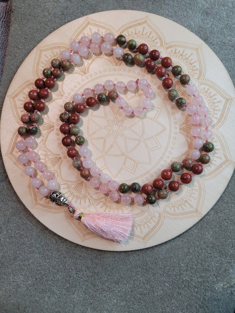 May include: A pink, green, and red gemstone mala with a silver tassel. The mala is on a wooden mandala.
