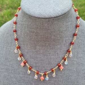 May include: A handmade necklace featuring red beads and gold wire. The necklace has small mushroom charms and iridescent crystal accents. The necklace is displayed on a gray mannequin head against a green background.
