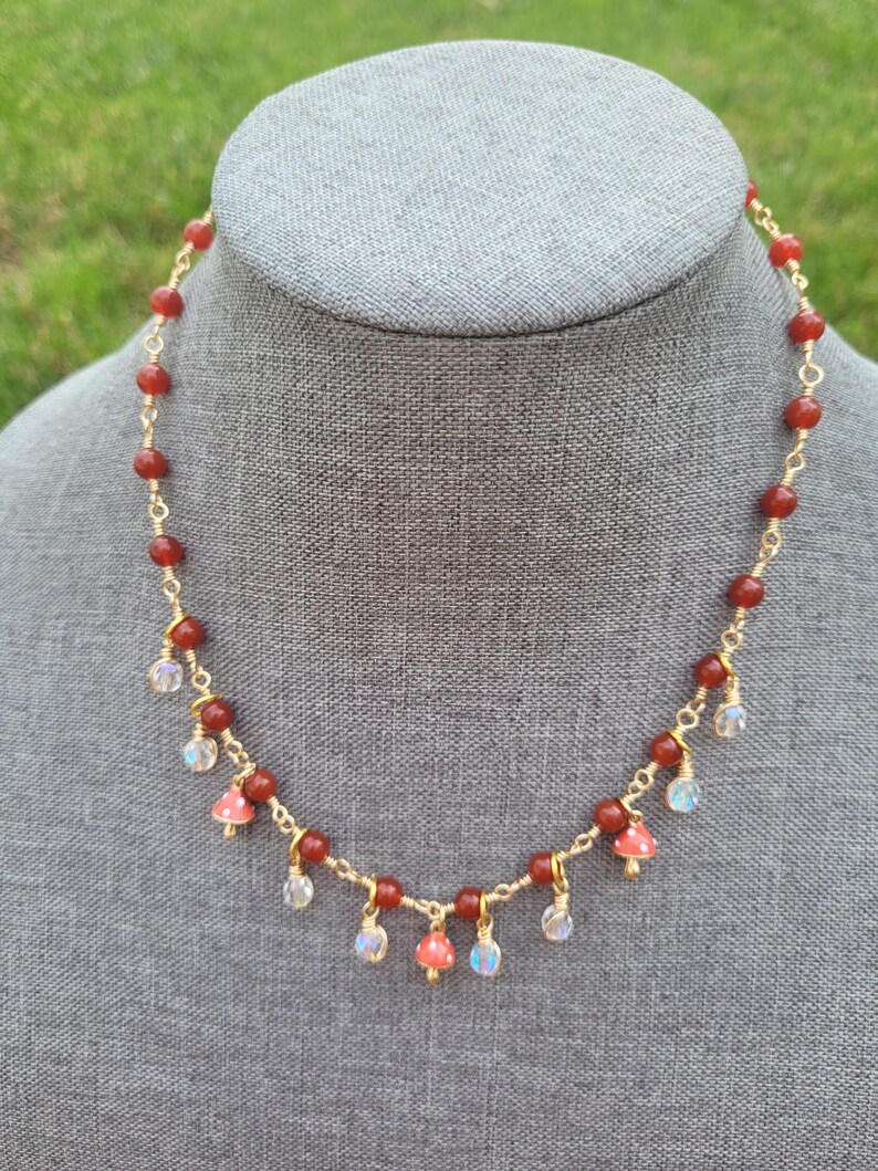 May include: A handmade necklace featuring red beads and gold wire. The necklace has small mushroom charms and iridescent crystal accents. The necklace is displayed on a gray mannequin head against a green background.
