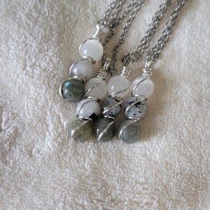 May include: Four necklaces with silver chains and wire-wrapped gemstone pendants. Each pendant features a combination of white, gray, and green spherical stones. The necklaces are displayed on a white, textured surface.