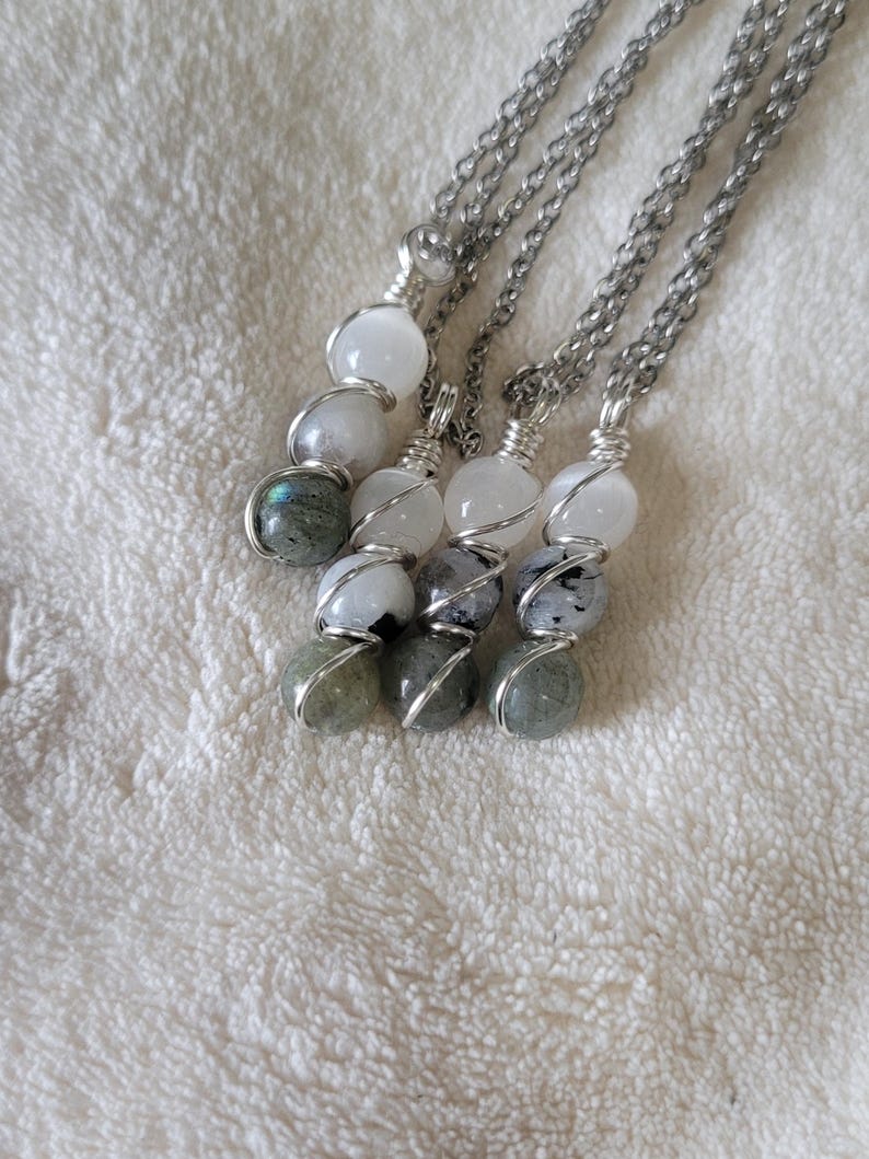 May include: Four necklaces with silver chains and wire-wrapped gemstone pendants. Each pendant features a combination of white, gray, and green spherical stones. The necklaces are displayed on a white, textured surface.