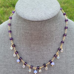 May include: A delicate necklace featuring amethyst beads and small, dangling charms. The necklace has a gold-colored chain and is displayed on a gray mannequin head. The amethyst beads are a deep purple color, and the charms are clear and iridescent.