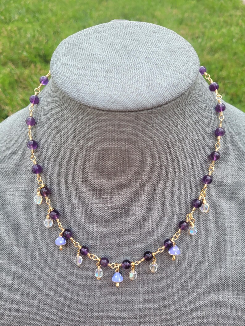 May include: A delicate necklace featuring amethyst beads and small, dangling charms. The necklace has a gold-colored chain and is displayed on a gray mannequin head. The amethyst beads are a deep purple color, and the charms are clear and iridescent.