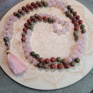 May include: A multi-strand mala necklace with pink, green, and brown beads. The necklace has a tassel at the end and is lying on a wooden surface with a mandala design.