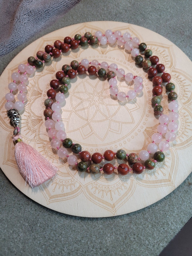 May include: A multi-strand mala necklace with pink, green, and brown beads. The necklace has a tassel at the end and is lying on a wooden surface with a mandala design.