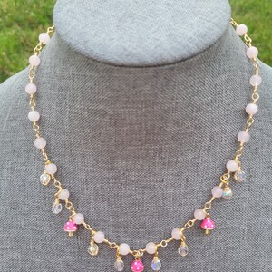 May include: A delicate necklace featuring pink gemstone beads and gold wire. The necklace has small, pink mushroom charms with white polka dots, and clear crystal accents. The necklace is displayed on a gray mannequin head.