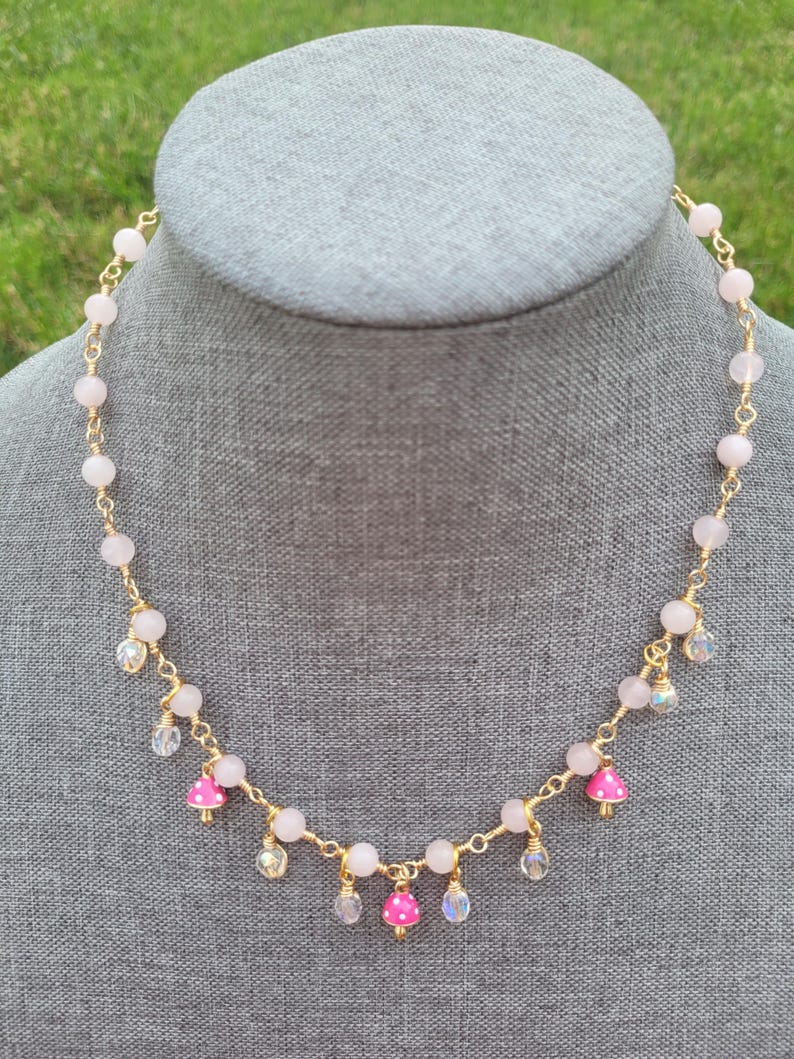 May include: A delicate necklace featuring pink gemstone beads and gold wire. The necklace has small, pink mushroom charms with white polka dots, and clear crystal accents. The necklace is displayed on a gray mannequin head.