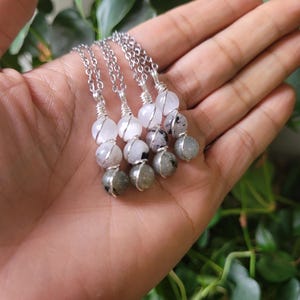 May include: Four silver necklaces with gemstone pendants. Each necklace features a chain and three beads: a white, a clear, and a gray stone. The stones are wire-wrapped together. The necklaces are displayed on a hand.
