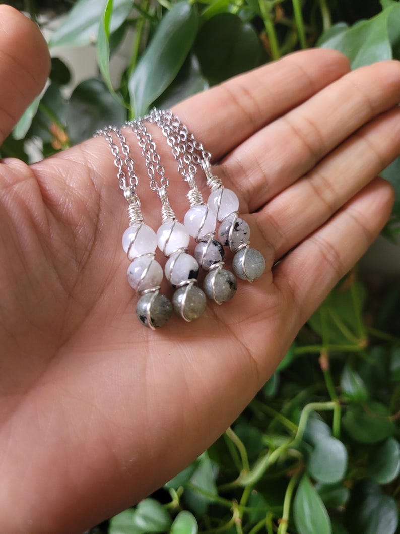May include: Four silver necklaces with gemstone pendants. Each necklace features a chain and three beads: a white, a clear, and a gray stone. The stones are wire-wrapped together. The necklaces are displayed on a hand.