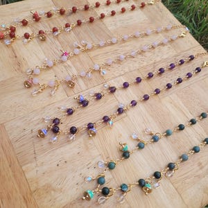 May include: Several beaded necklaces with gold chains and various gemstone beads, including red, pink, purple, and green. Each necklace has small charms and dangles. The necklaces are displayed on a wooden surface.