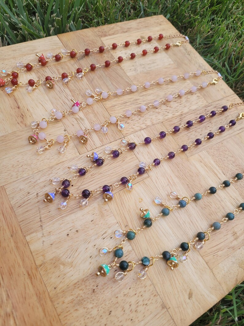May include: Several beaded necklaces with gold chains and various gemstone beads, including red, pink, purple, and green. Each necklace has small charms and dangles. The necklaces are displayed on a wooden surface.