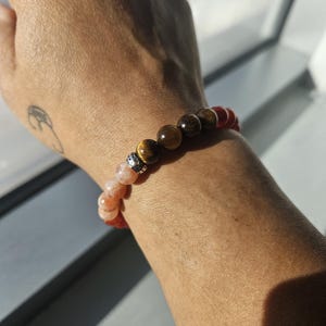 May include: A beaded bracelet with a silver charm, featuring orange, brown, red, and pink stones.