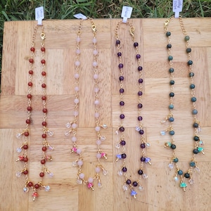May include: Four beaded necklaces with gold chains and accents. The necklaces feature various colored beads, including red, pink, purple, and green. Each necklace has a different style of dangling charms and is displayed on a wooden surface.