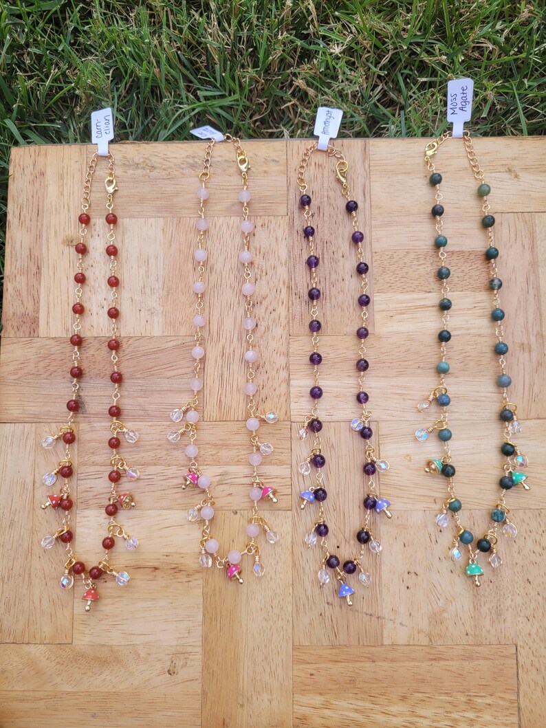 May include: Four beaded necklaces with gold chains and accents. The necklaces feature various colored beads, including red, pink, purple, and green. Each necklace has a different style of dangling charms and is displayed on a wooden surface.