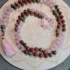 May include: A mala or prayer beads made with three different types of stones. The mala is made with pink rose quartz, brown red jasper, and green moss agate stones. The mala has a pink tassel.