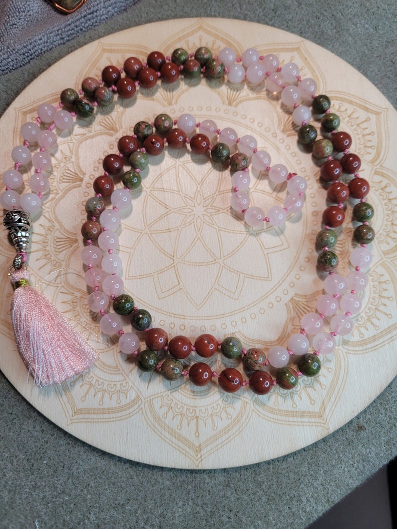 May include: A mala or prayer beads made with three different types of stones. The mala is made with pink rose quartz, brown red jasper, and green moss agate stones. The mala has a pink tassel.