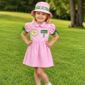 May include: A light pink dress with short puff sleeves featuring green ribbon ties, a collared neckline, and a front zipper. The chest displays a circular Caddie Future Pro patch and a green rectangular patch with the name Emma and the number 1.