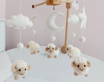 May include: A wooden baby mobile with white felt clouds, a crescent moon, and stars. There are five crocheted white sheep with brown faces hanging from the mobile. The mobile is hanging from a white ceiling.
