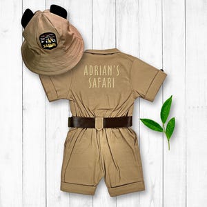 May include: A tan safari jumpsuit with a brown belt and short sleeves. The jumpsuit has "Adrian's Safari" embroidered on the back. A tan safari hat with black ears is included.