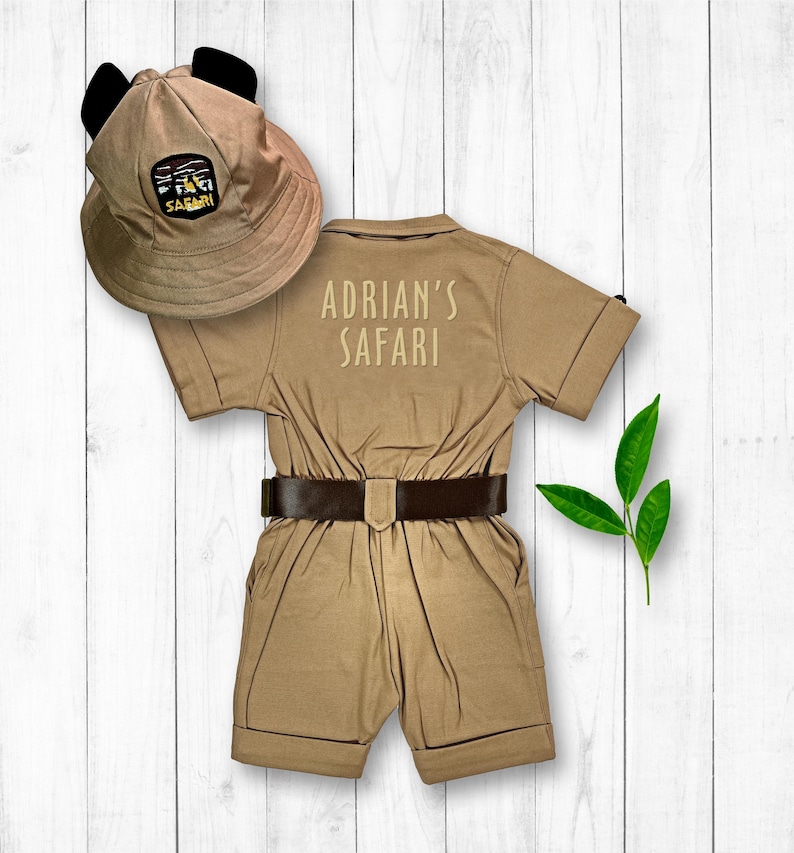 May include: A tan safari jumpsuit with a brown belt and short sleeves. The jumpsuit has "Adrian's Safari" embroidered on the back. A tan safari hat with black ears is included.