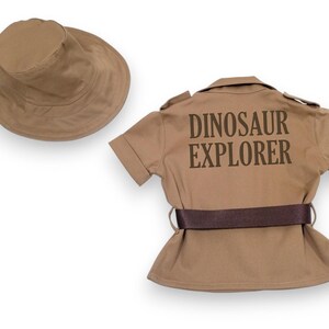 Puede incluir: Un conjunto de disfraz infantil de color beige, que incluye un sombrero de ala ancha y una camisa de manga corta con el texto "DINOSAUR EXPLORER" impreso en la espalda. La camisa tiene un cintur&oacute;n marr&oacute;n.