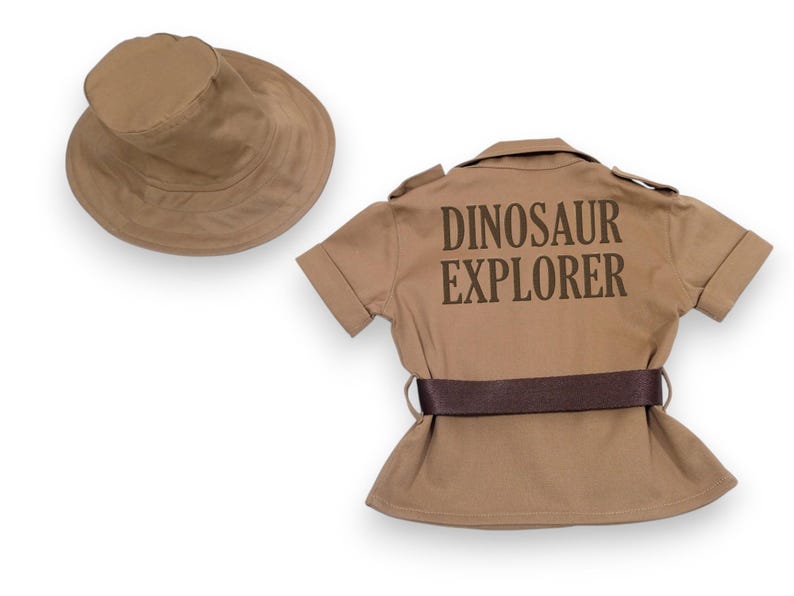 Puede incluir: Un conjunto de disfraz infantil de color beige, que incluye un sombrero de ala ancha y una camisa de manga corta con el texto "DINOSAUR EXPLORER" impreso en la espalda. La camisa tiene un cintur&oacute;n marr&oacute;n.