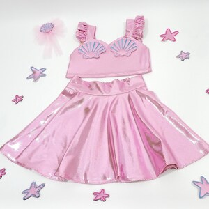 May include: A pink two-piece outfit for girls, featuring a sparkly skirt and a crop top with a seashell design.
