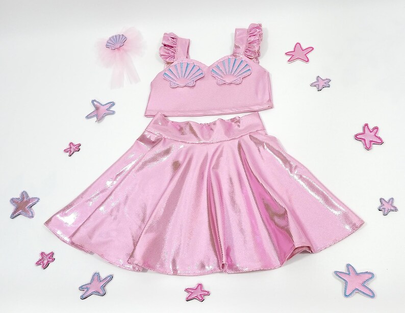 May include: A pink two-piece outfit for girls, featuring a sparkly skirt and a crop top with a seashell design.